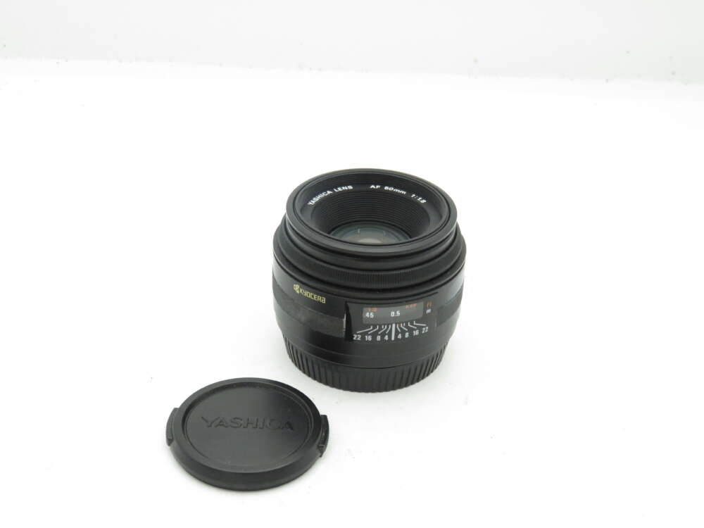 IMG_0654 Camera lens marked "Yashica Lens AF 50mm 1:1.8" with a Yashica lens cap beside it.