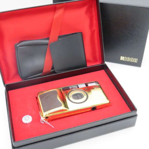 Gold Ricoh camera with case, presented in a black box lined with red fabric.