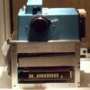 A vintage, large-format camera with a metallic base and blue top housing, featuring prominent knobs and dials.
