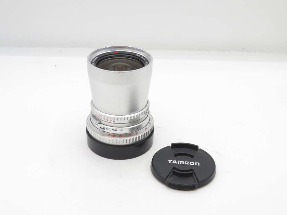 IMG_1202 Silver camera lens with black "Tamron" lens cap placed beside it on a white background.