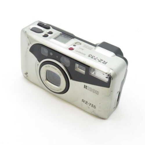 A silver and black Ricoh RZ-735 point-and-shoot film camera featuring a built-in lens, a viewfinder, and various buttons and switches on the top and back. The camera appears to have signs of wear and use with some scratches visible on its body.