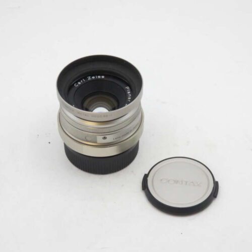 A Carl Zeiss Planar camera lens with a silver Contax lens cap beside it on a white surface.