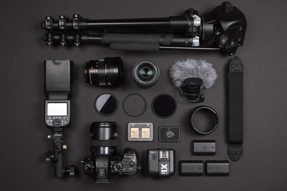 Professional camera equipment including a tripod, lenses, camera, microphone, flash, battery pack, filters, memory cards, and camera strap arranged on a dark surface.
