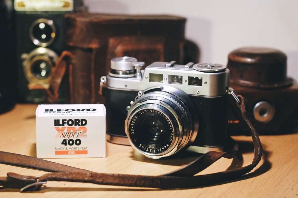 Vintage rangefinder camera with a leather strap beside a box of Ilford XP2 Super 400 black and white film on a wooden surface, with old camera accessories in the background.