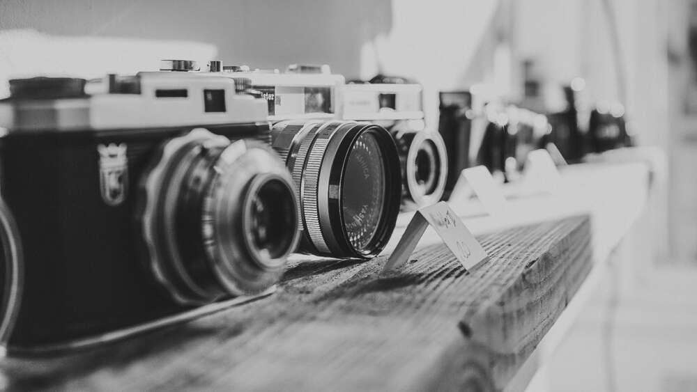 Black and white photo of vintage cameras displayed on a wooden shelf.