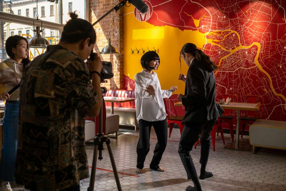 A film crew shoots a scene inside a restaurant with a vibrant map mural on the wall; a camera operator focuses on two people performing a dramatic confrontation.