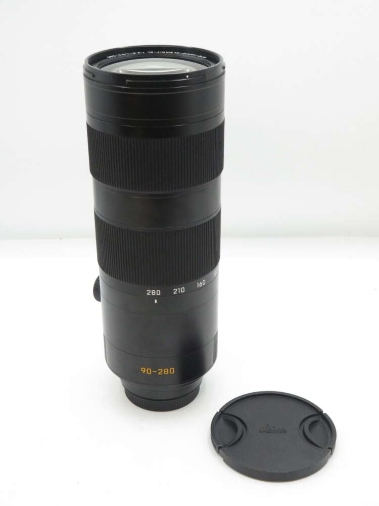 IMG_2880 Black camera lens with a 90-280mm zoom range, placed upright with a detached lens cap beside it.