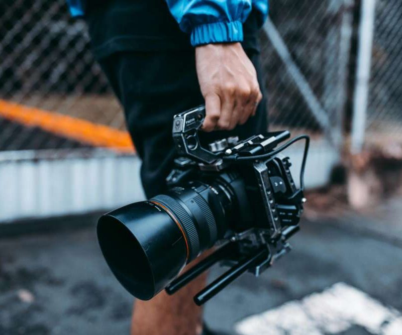 Person holding a professional camera by their side, with a blurred background of a fence and pavement.