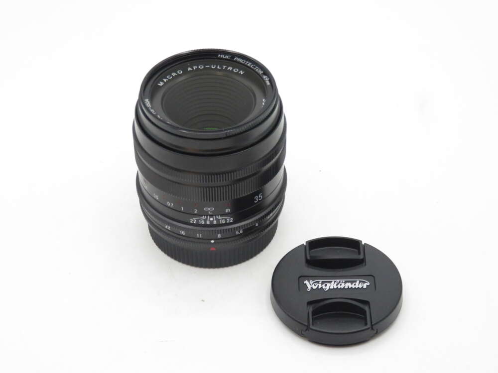 IMG_0349 A black Voigtländer camera lens labeled 'MACRO APO-ULTRON' with focus and aperture adjustment rings, next to a matching lens cap on a plain white background.