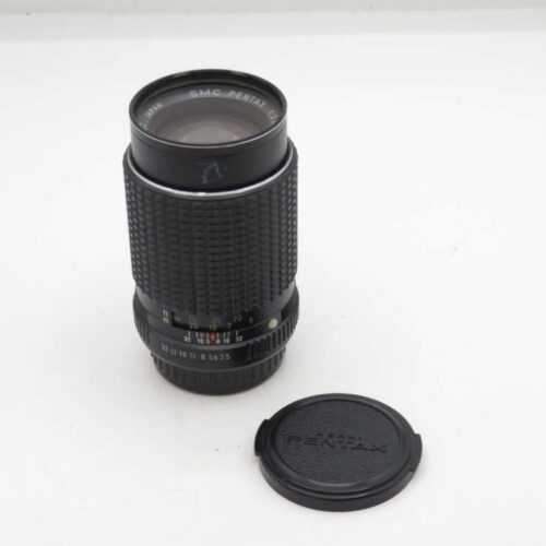 A black SMC Pentax camera lens with its detached lens cap placed beside it on a white surface.