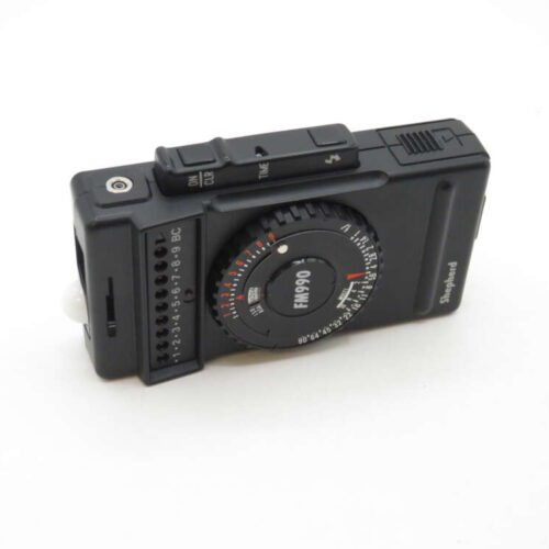 Black analog light meter with a circular dial and slider switch, labeled FM990 and Shepherd, on a white background.