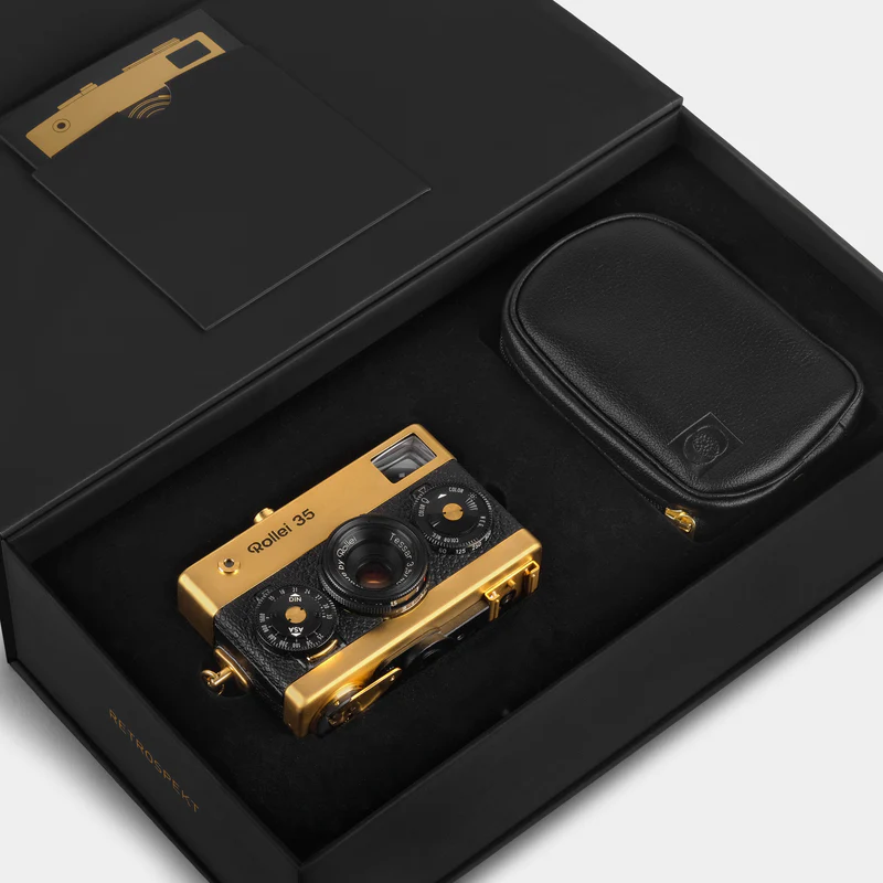 A gold and black Rollei 35 camera displayed in an open black box with a black leather pouch beside it.