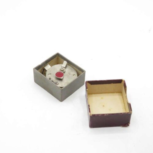 A small metal mechanical device with a red button and levers, housed in an open square box with a separate lid placed nearby, on a white background.