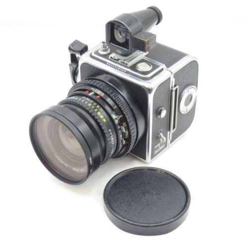 A vintage medium format camera with a large lens, angular design, and a viewfinder on top, resting on a white surface beside a detached lens cap.