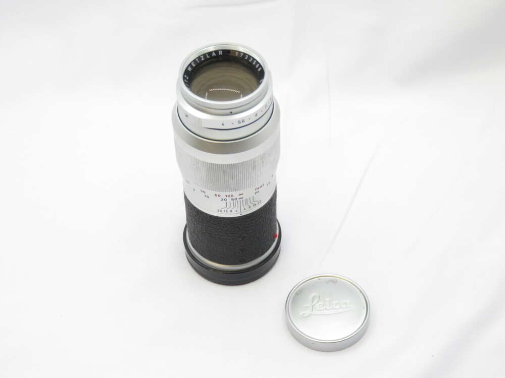 IMG_0504 A vintage Leica camera lens with a silver and black body, featuring aperture and focus markings. The lens cap is placed next to it on a white surface.
