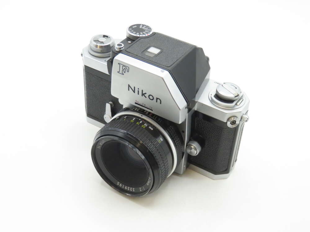 IMG_0551 A vintage Nikon F SLR camera with a black and silver body and a lens attached, resting on a white surface.