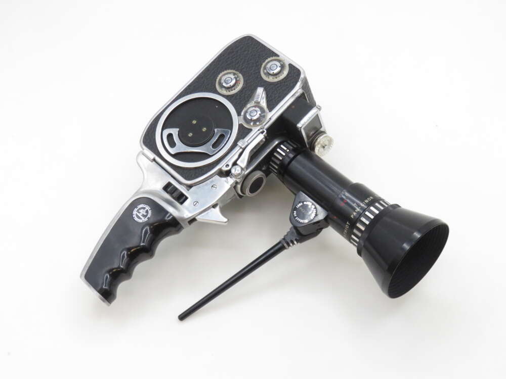 A vintage handheld film camera with a black grip handle and a long black lens attachment, featuring silver metal accents and a wheel dial on a plain white background.