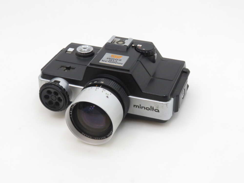 IMG_0623 A vintage Minolta Minoltina-P 8mm movie camera with a black and silver body, featuring a prominent lens and dials for adjustments, set against a plain white background.