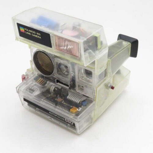 A Polaroid 660 Land Camera with a transparent casing, revealing its internal components and circuitry, set against a plain white background.