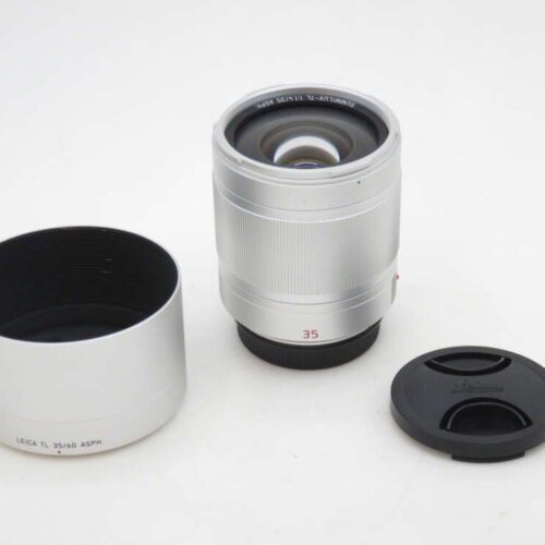 A silver Leica TL 35mm f/1.4 ASPH lens with its lens hood and lens cap on a white background.