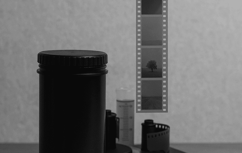 A black and white image of a film developing scene, featuring a roll of film strip hanging in front of a large black film canister. In the background, a film developing tank and a partially unspooled roll of film are placed on a surface.