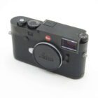 A black Leica camera with a lens cap, featuring a red Leica logo on the front and manual dials on top, set against a plain white background.