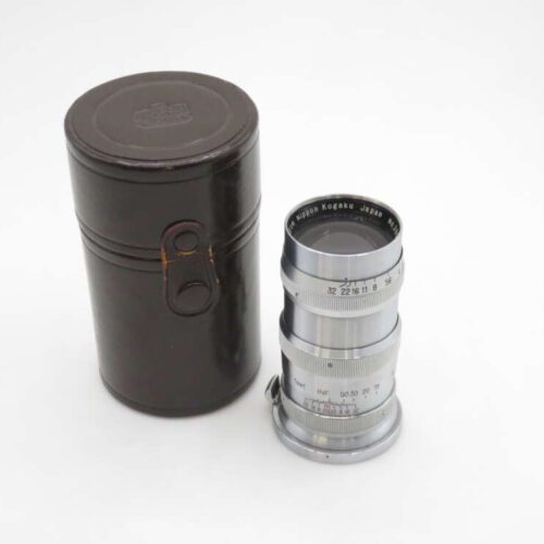 A vintage camera lens labeled "Nippon Kogaku Japan" next to its cylindrical black case on a white background.