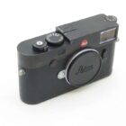 A black Leica camera with various control dials and a lens cap displaying the Leica logo, placed on a white surface.