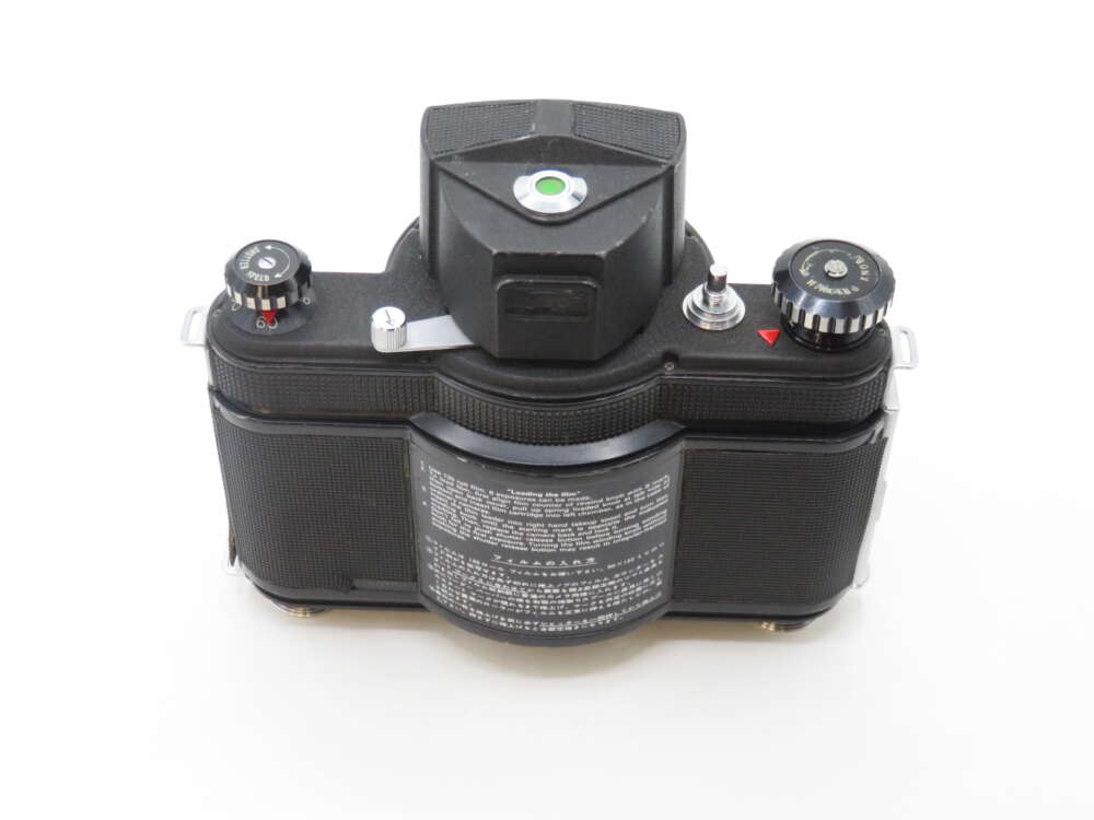 A vintage black camera viewed from the top, showing dials and a rectangular viewfinder, with instructional text in white on the back plate.