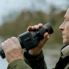 A person outdoors using a monocular device for viewing distant objects, wearing a green jacket with a light-coloured collar, against a blurred natural background.