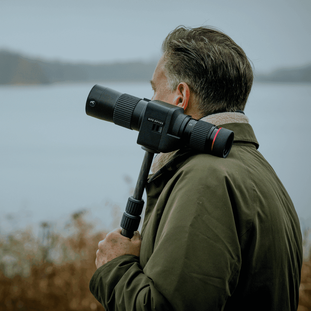 A person wearing a green jacket holding a telescope with a tripod near a body of water, focusing on the view ahead.