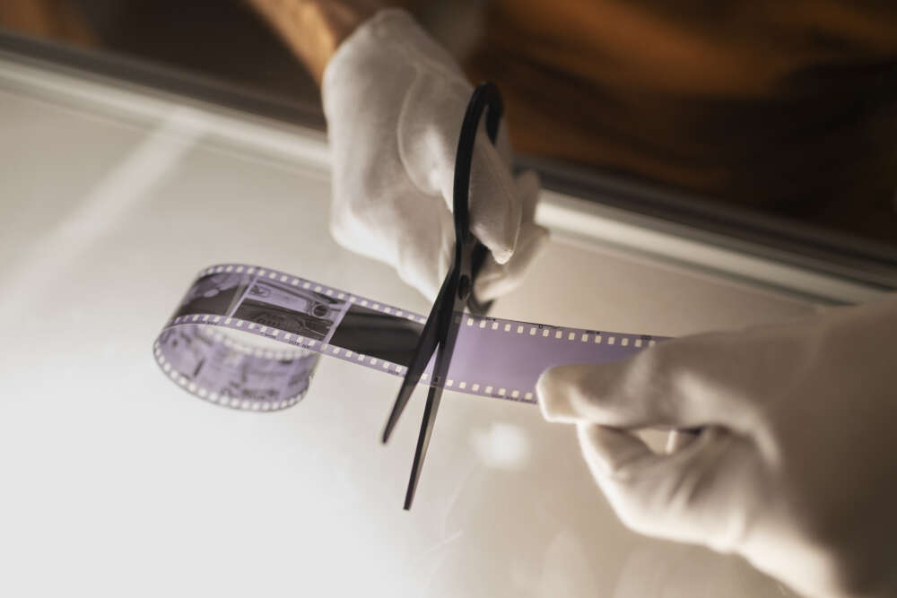 A person wearing white gloves is cutting a strip of photographic film with scissors on a backlit surface.