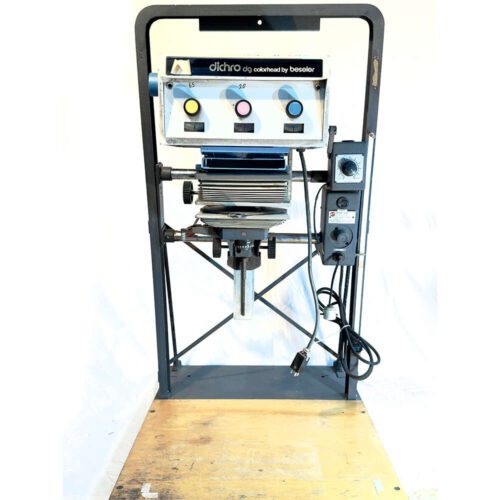 An enlarger with a Dichro colorhead by Beseler, featuring three dials for adjusting color settings, mounted on a stand with a plug cord visible, set against a plain backdrop.