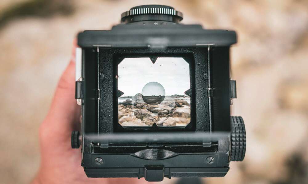 Close-up of a person's hand holding a vintage camera, focusing on a rocky landscape and sky visible through the camera's viewfinder.