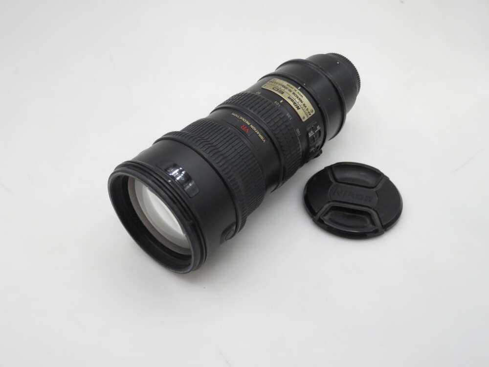 IMG_1147 A Nikon telephoto camera lens with a black body, resting on its side next to a detached lens cap, on a white background.