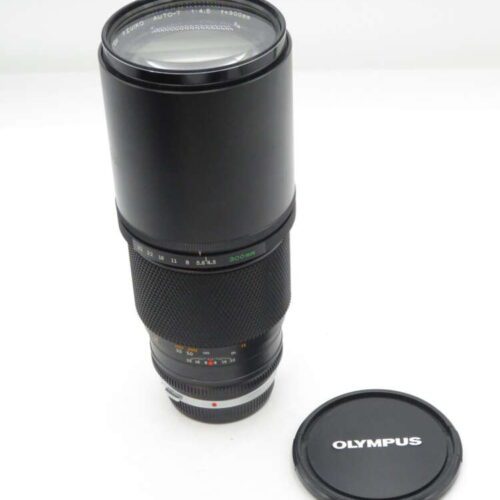A black Olympus camera lens with a lens cap next to it on a white background, featuring focal length markings and an aperture ring.