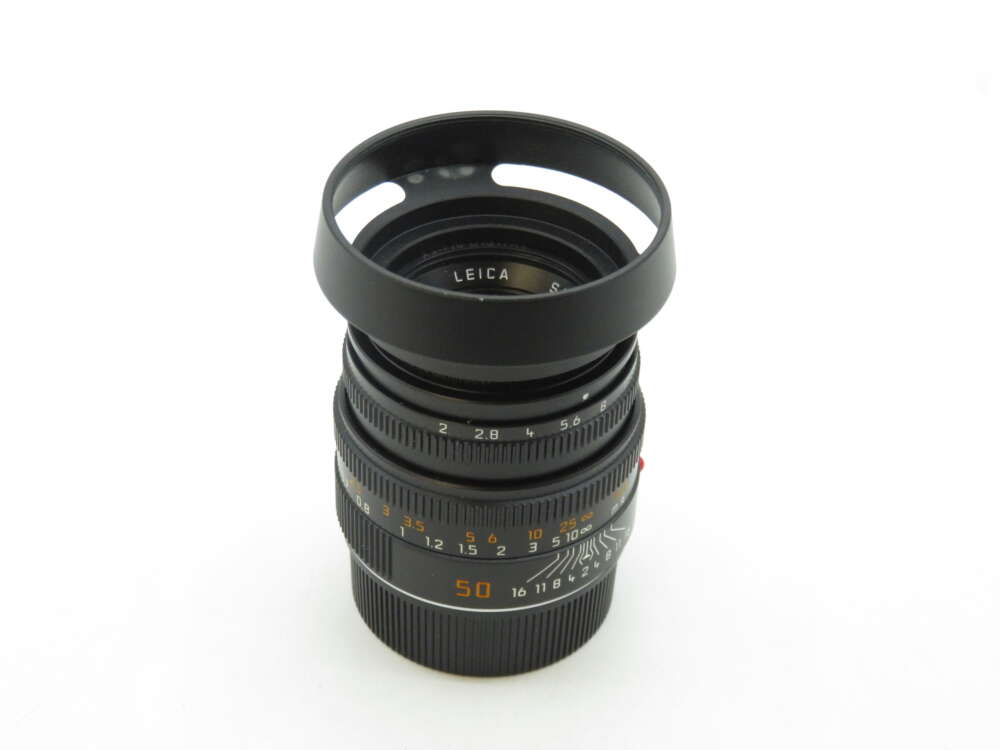 IMG_1333 A Leica camera lens with a lens hood attached, showing various aperture and focus markings on the barrel, set against a plain white background.