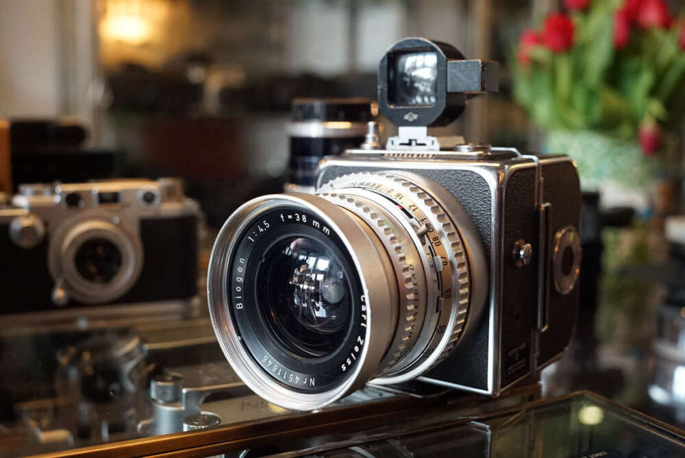 A vintage medium format camera with a large Carl Zeiss Biogon lens displayed on a glass surface, with other vintage cameras blurred in the background.