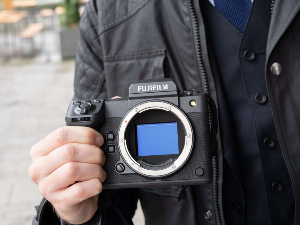 A person holding a Fujifilm camera body without a lens, displaying the sensor and various control buttons, against an outdoor background.