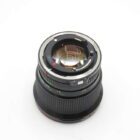 A camera lens with a metallic mount, viewed from the back on a white background. The markings and aperture blades are visible through the lens elements.
