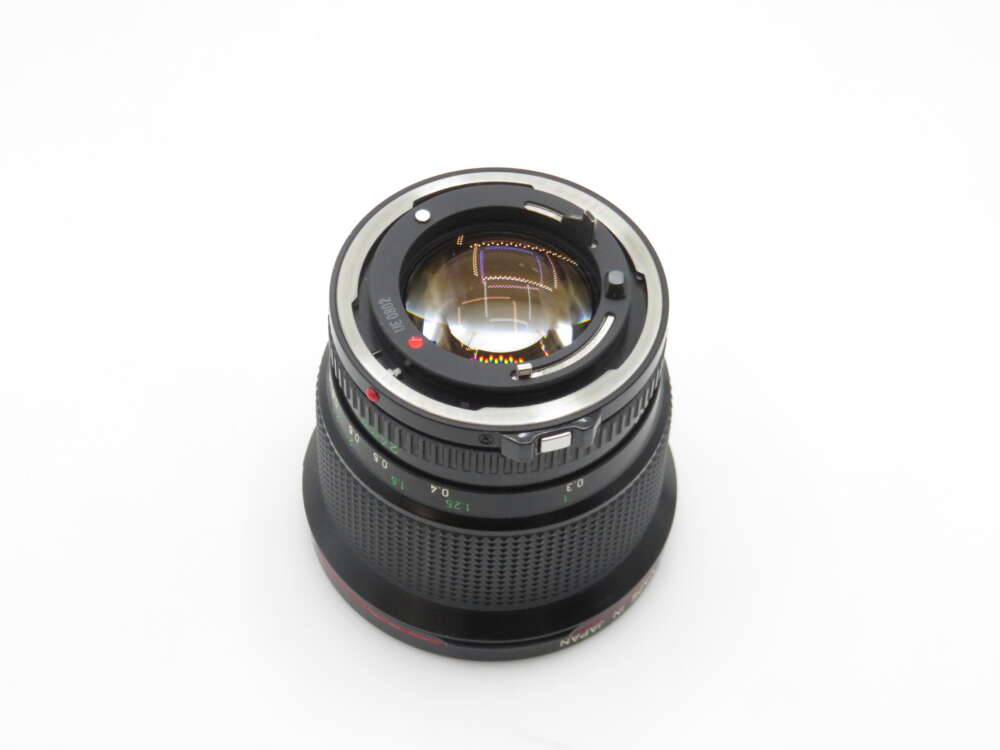 A camera lens with a metallic mount, viewed from the back on a white background. The markings and aperture blades are visible through the lens elements.