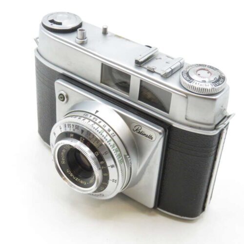 A vintage film camera with a silver finish and black leather-like covering, featuring a prominent lens with various settings and dials on top of the body, including a viewfinder and exposure meter.