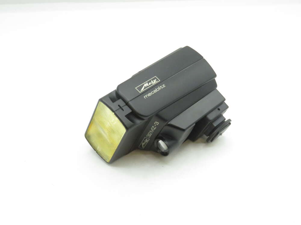 IMG_1691 A Metz mecablitz 32 MZ-3 camera flash unit rests on a white surface, featuring a black plastic body with a yellow-tinted diffuser at the front.