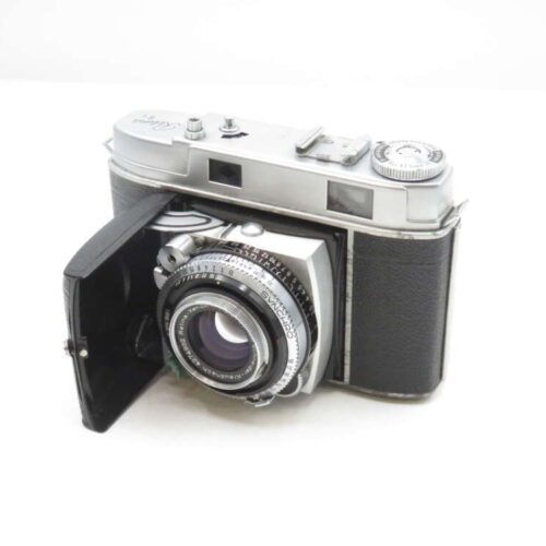A vintage film camera with a black leather covering and silver metallic accents, featuring a large lens and several manual controls. The camera's front panel is open, revealing the detailed lens mechanism.