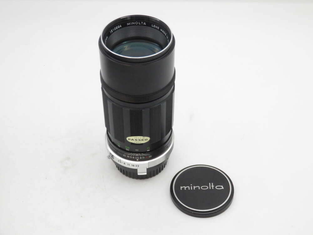 IMG_1703 A Minolta camera lens with a black body, silver mount, and an attached 'PASSED' sticker, positioned beside a Minolta lens cap on a white background.