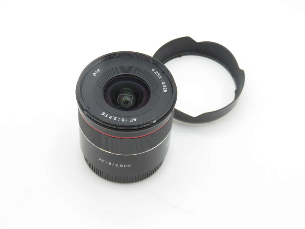IMG_1717 A camera lens with the label 'AF 18/2.8 FE' positioned next to a lens hood on a plain white background.
