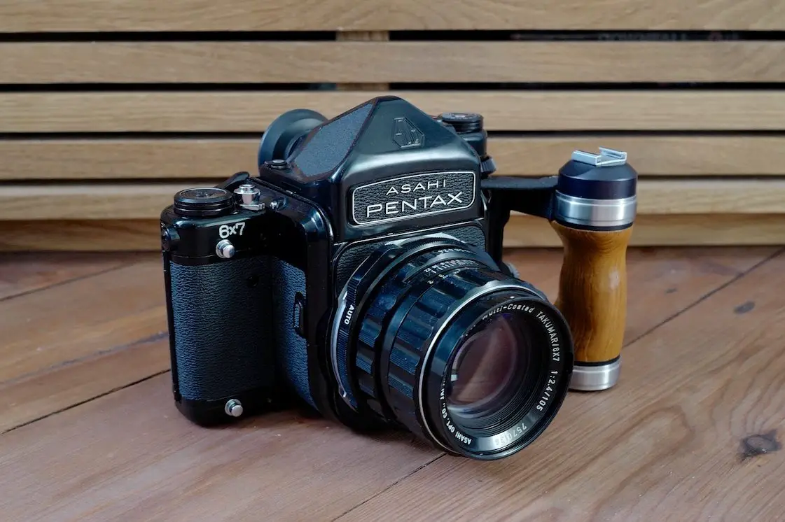 A black Asahi Pentax 6x7 medium format film camera with a wooden grip, placed on a wooden surface with a wooden background. The camera features a large lens and has a vintage design.