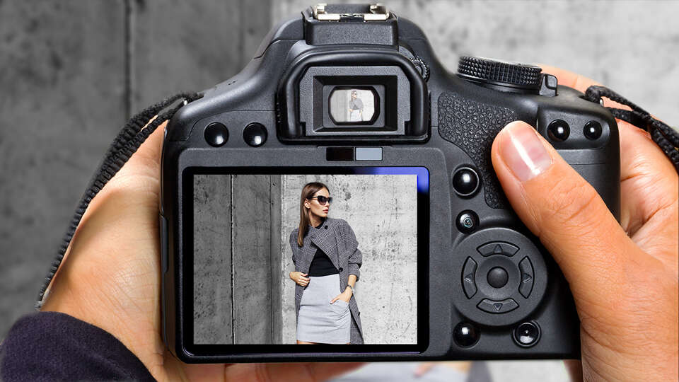 A digital camera displaying an image of a woman posing against a concrete wall, wearing sunglasses, a grey sweater, and a light-coloured skirt, with her hands in her pockets.