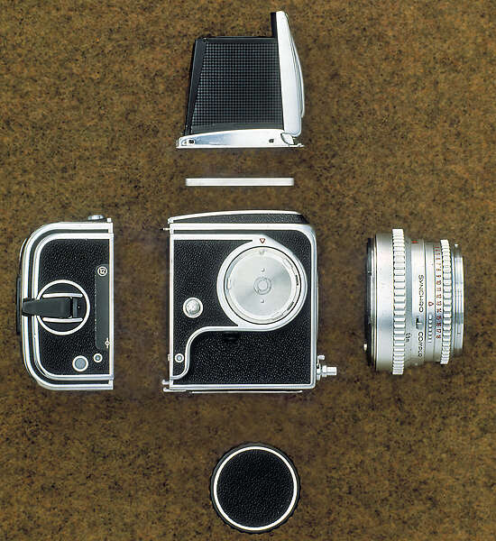 Disassembled camera with separate components including a lens, viewfinder, body, film back, and cap, arranged on a brown textured surface.