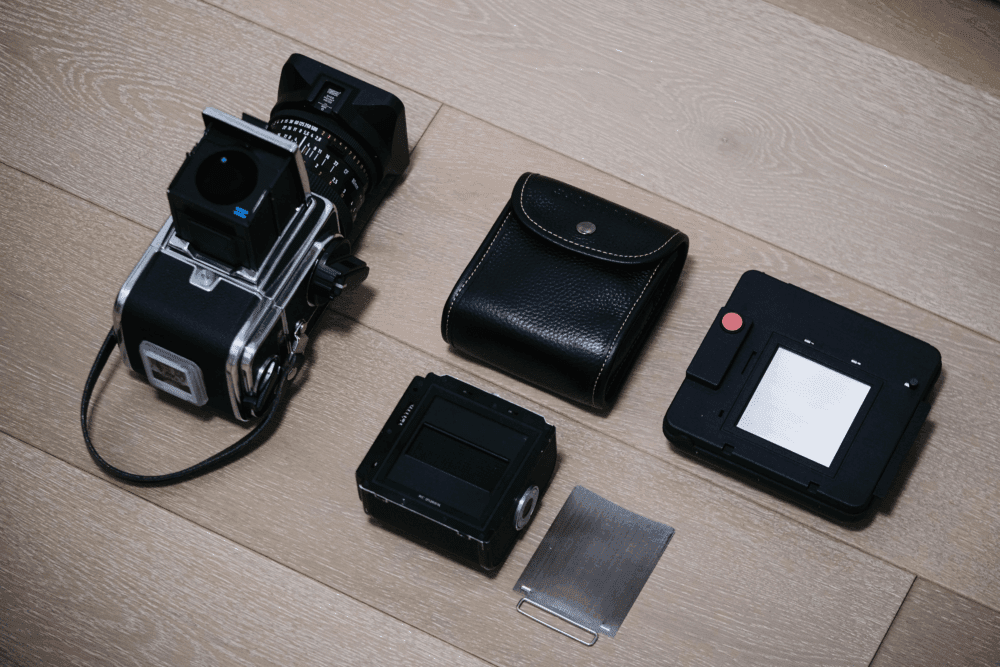 A set of camera equipment on a wooden floor, including a vintage medium format camera with an attached lens, a film magazine, a black pouch, a camera back with a red button, and a metal slide component.